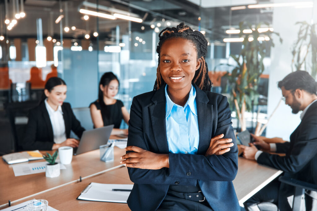 A young professional standing confidently in a modern office setting with her team brainstorming in the background
